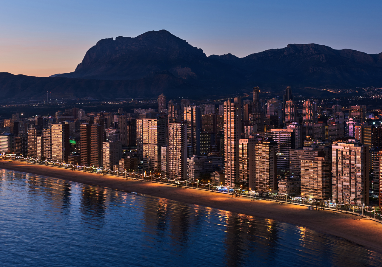Una plaza urbana soleada con edificios blancos ornamentados, una gran fuente y personas en la calle.