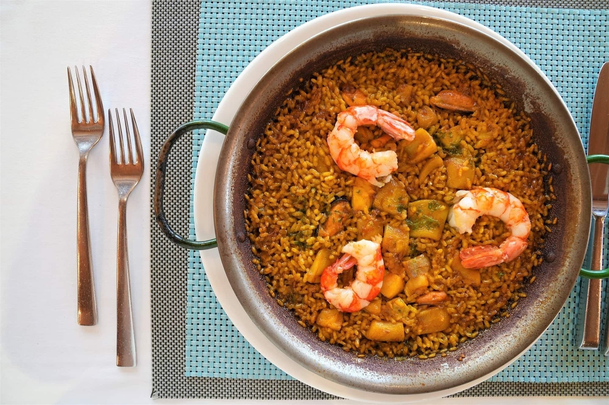 a pan filled with rice and shrimp sits on a table next to forks and knives