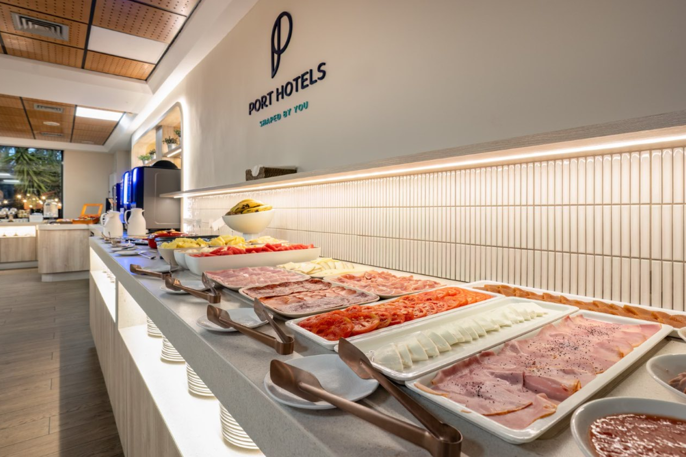 La imagen muestra un moderno buffet de desayuno de Port Hotels, con una amplia variedad de fiambres, quesos y frutas frescas.