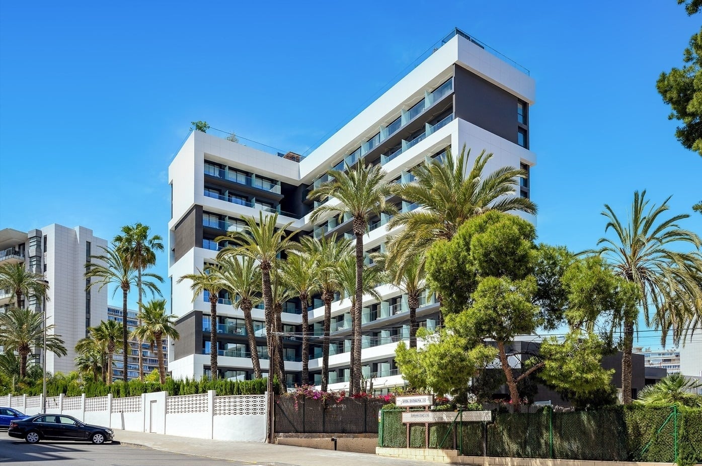 Modernos edificios blancos y grises se alzan entre exuberantes palmeras bajo un cielo azul claro, con una calle y un coche en el primer plano.
