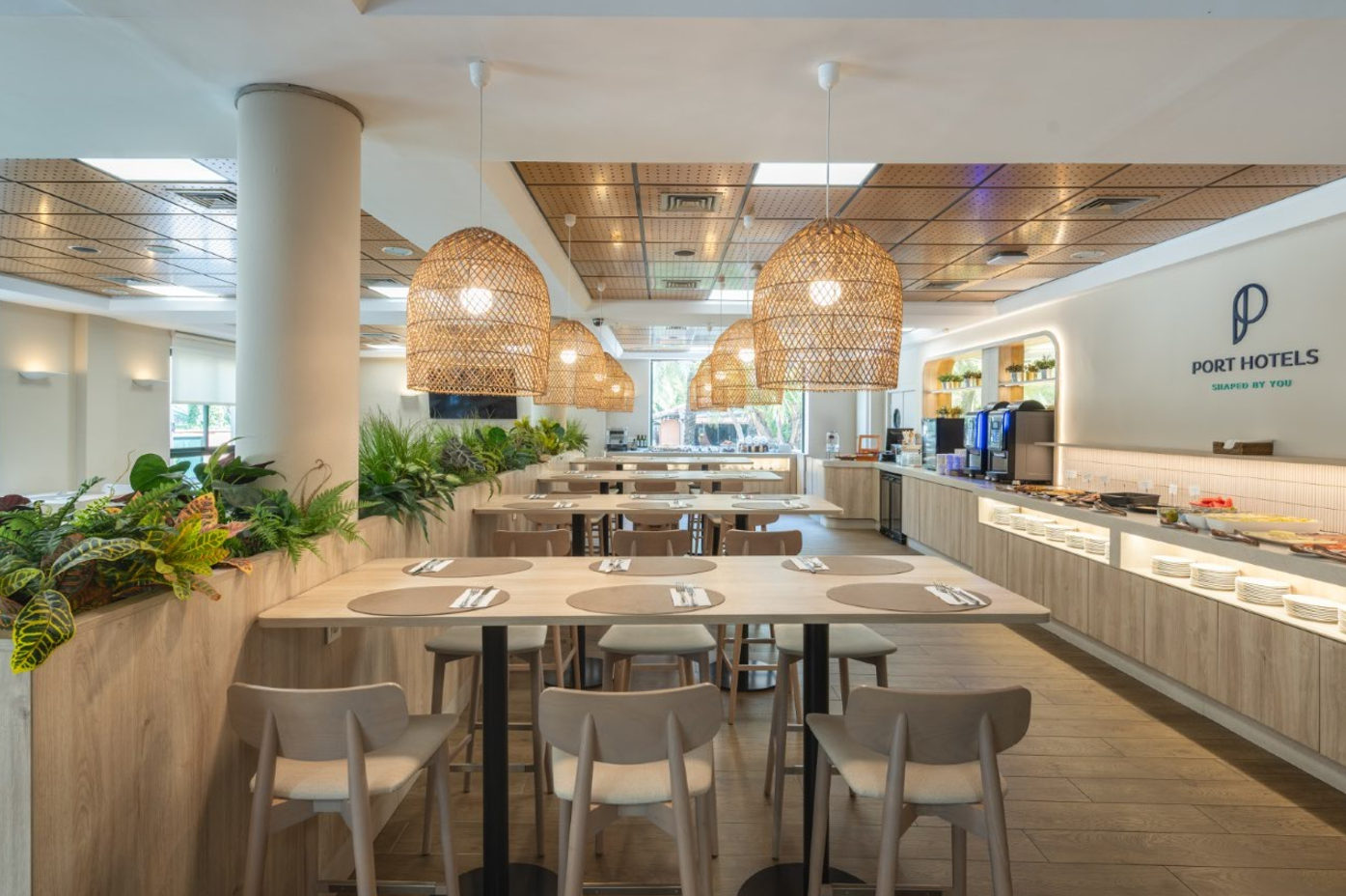 Un restaurante moderno y luminoso, decorado con muebles de madera clara, lámparas de mimbre y plantas, presenta un área de buffet con diversas estaciones de comida.