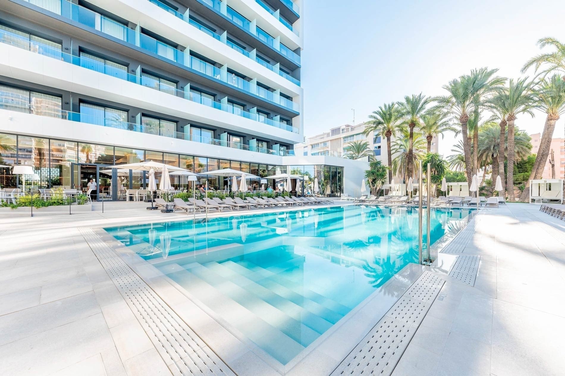 Un moderno hotel con una gran piscina al aire libre, rodeada de tumbonas y palmeras, bajo un cielo azul.