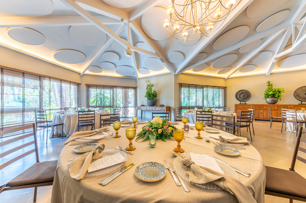 Una sala de reuniones luminosa y moderna está equipada con mesas vestidas, sillas, botellas de agua y cuadernos, con ventanas circulares que dan a un entorno natural de palmeras.