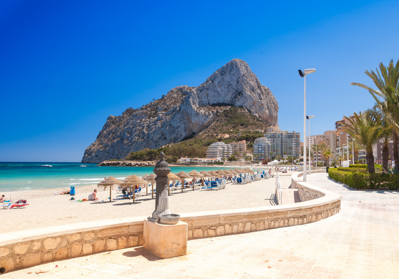 Panorámica de una ciudad costera densamente edificada con un puerto deportivo y montañas al fondo, bañada por aguas azules bajo un cielo despejado.