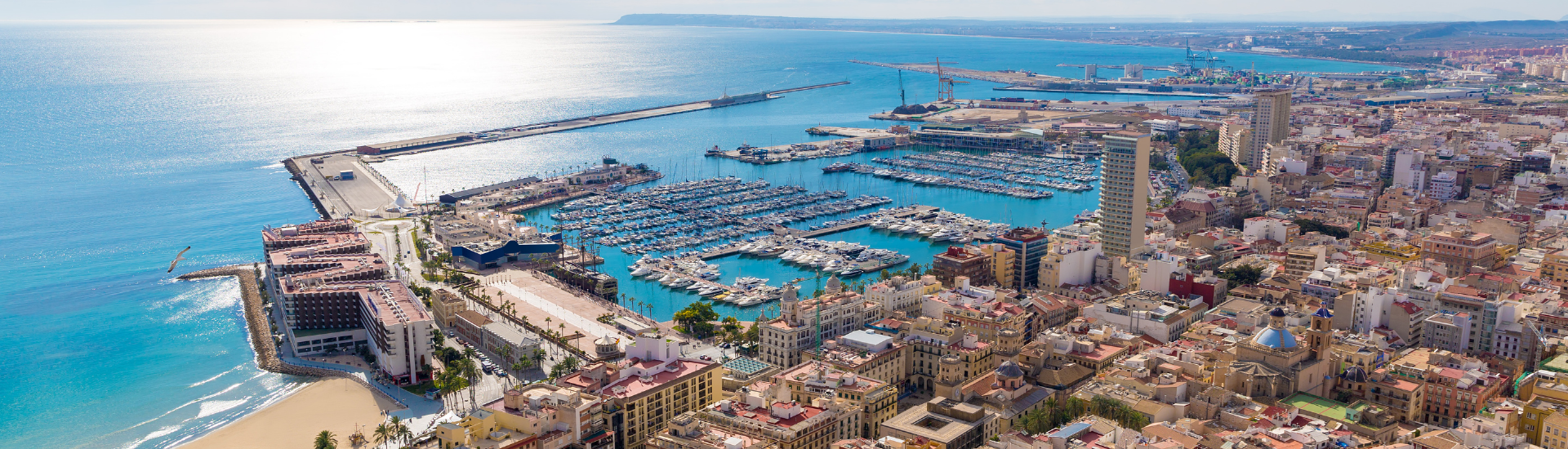 La imagen muestra una panorámica costera con un imponente peñón rocoso, aguas marinas de un azul intenso con varias embarcaciones, una playa con edificios y un puerto.