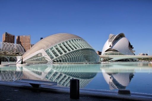 La moderna y futurista arquitectura de la Ciudad de las Artes y las Ciencias de Valencia se refleja en una lámina de agua bajo un cielo azul despejado.