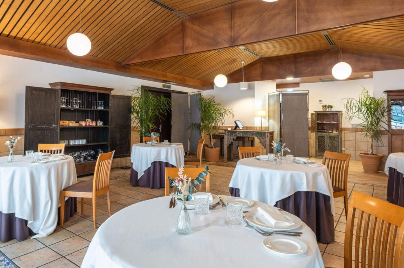 Un acogedor comedor de restaurante presenta mesas redondas con manteles blancos y bases oscuras, sillas de madera, un techo de vigas de madera, plantas decorativas y lámparas colgantes esféricas.