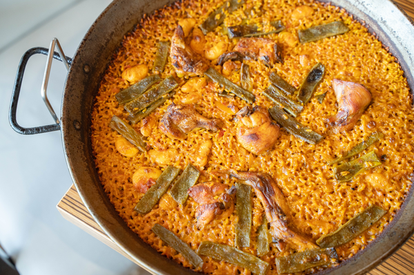 El interior de un restaurante con mesas puestas, destacando platos rojos y blancos, vasos, servilletas y un menú azul en primer plano, con vigas de madera en el techo.
