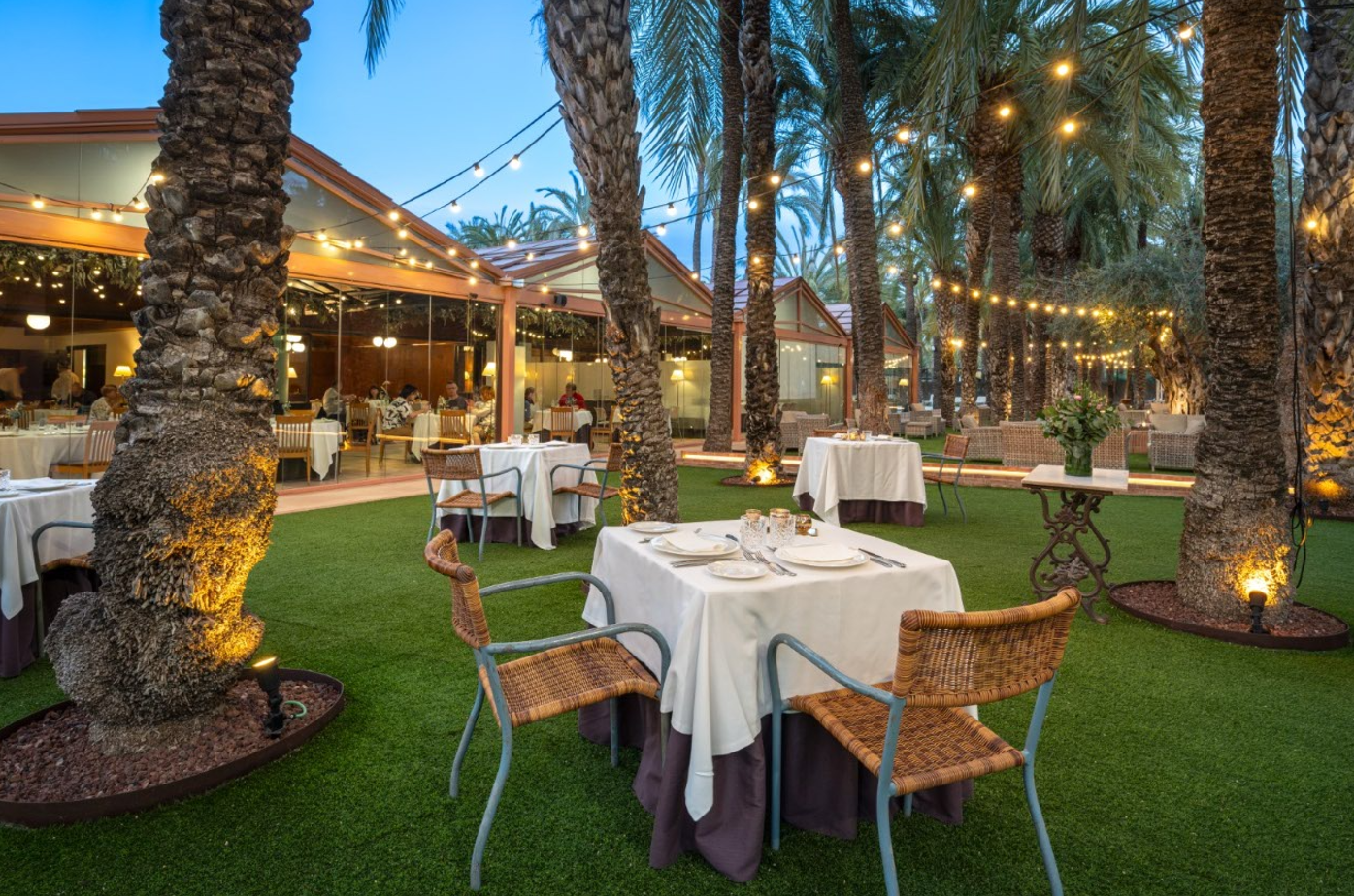 Un elegante restaurante al aire libre presenta mesas dispuestas sobre césped entre palmeras iluminadas con guirnaldas de luces, y un comedor acristalado con comensales al fondo.