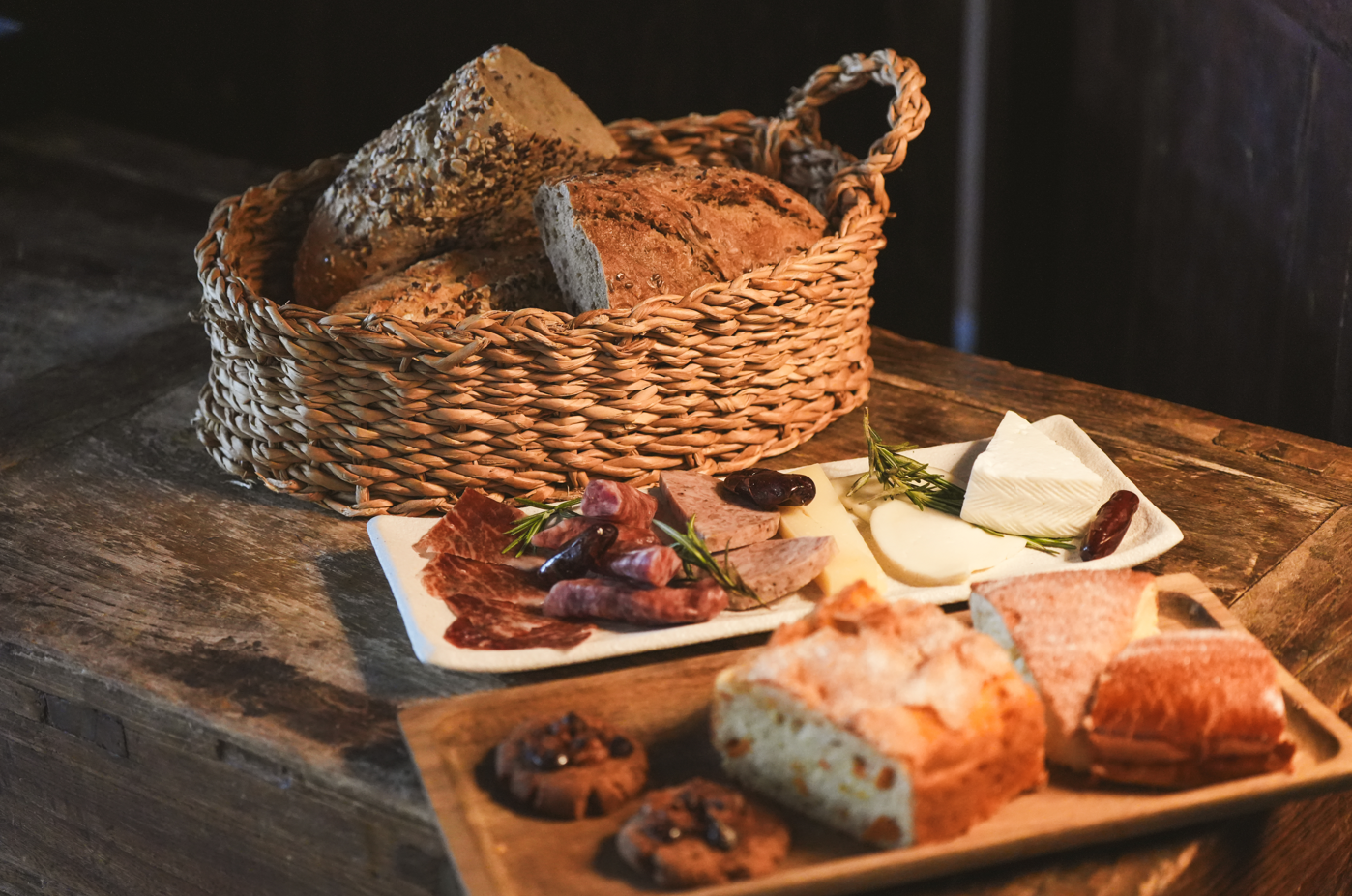 Una mesa de madera rústica presenta una cesta de pan, una tabla con embutidos, quesos y dátiles, y una bandeja con más pan y galletas.