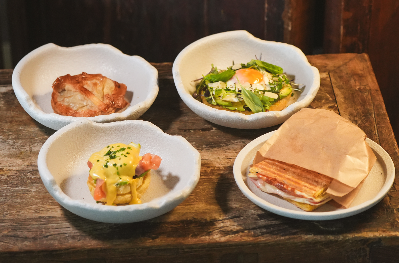 Cuatro pequeños platos con variadas preparaciones culinarias, que incluyen una tostada con huevo, huevos Benedict, un pastel y un sándwich, están dispuestos sobre una superficie de madera.