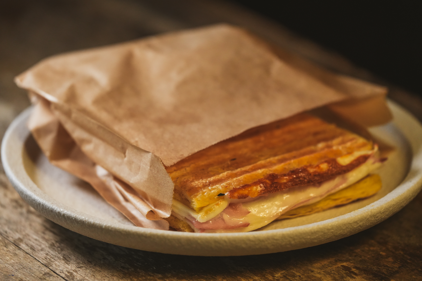 Un sándwich tostado de jamón y queso derretido, parcialmente envuelto en papel marrón, se encuentra en un plato sobre una superficie de madera.