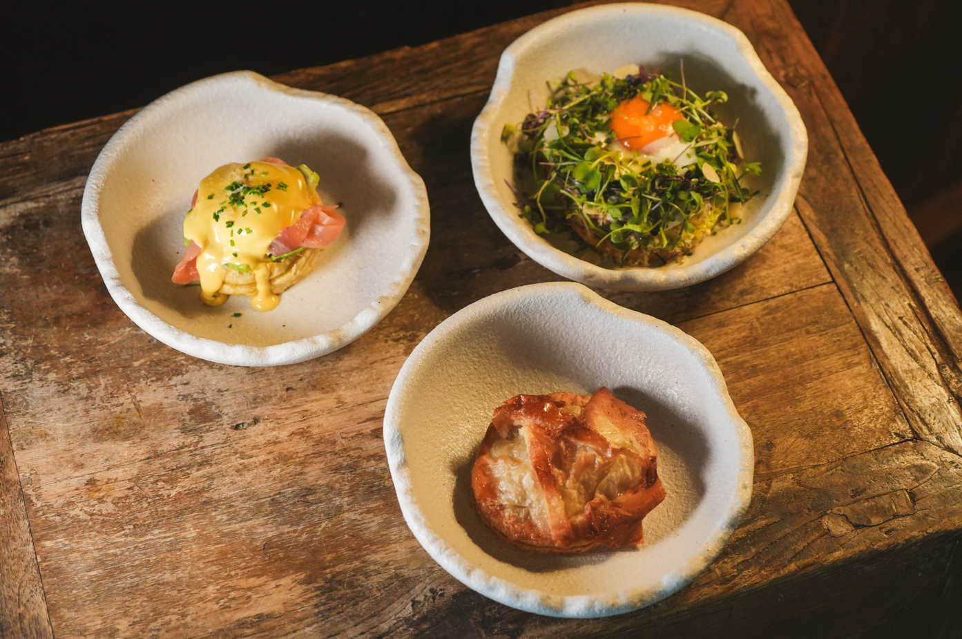 es: Tres cuencos de cerámica blanca que contienen un huevo Benedict con salmón, una ensalada de microgreens con yema de huevo y un hojaldre, se presentan sobre una mesa de madera rústica.