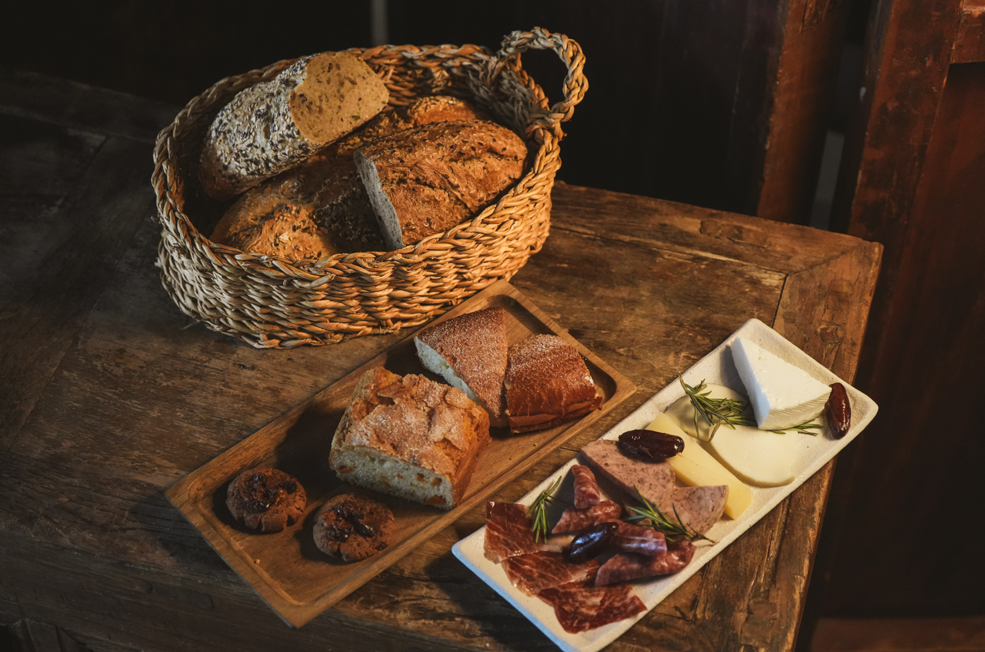 Una mesa de madera rústica presenta una cesta de pan artesanal, una bandeja con pan y galletas, y un plato con quesos, embutidos y dátiles, todo bajo una iluminación cálida.