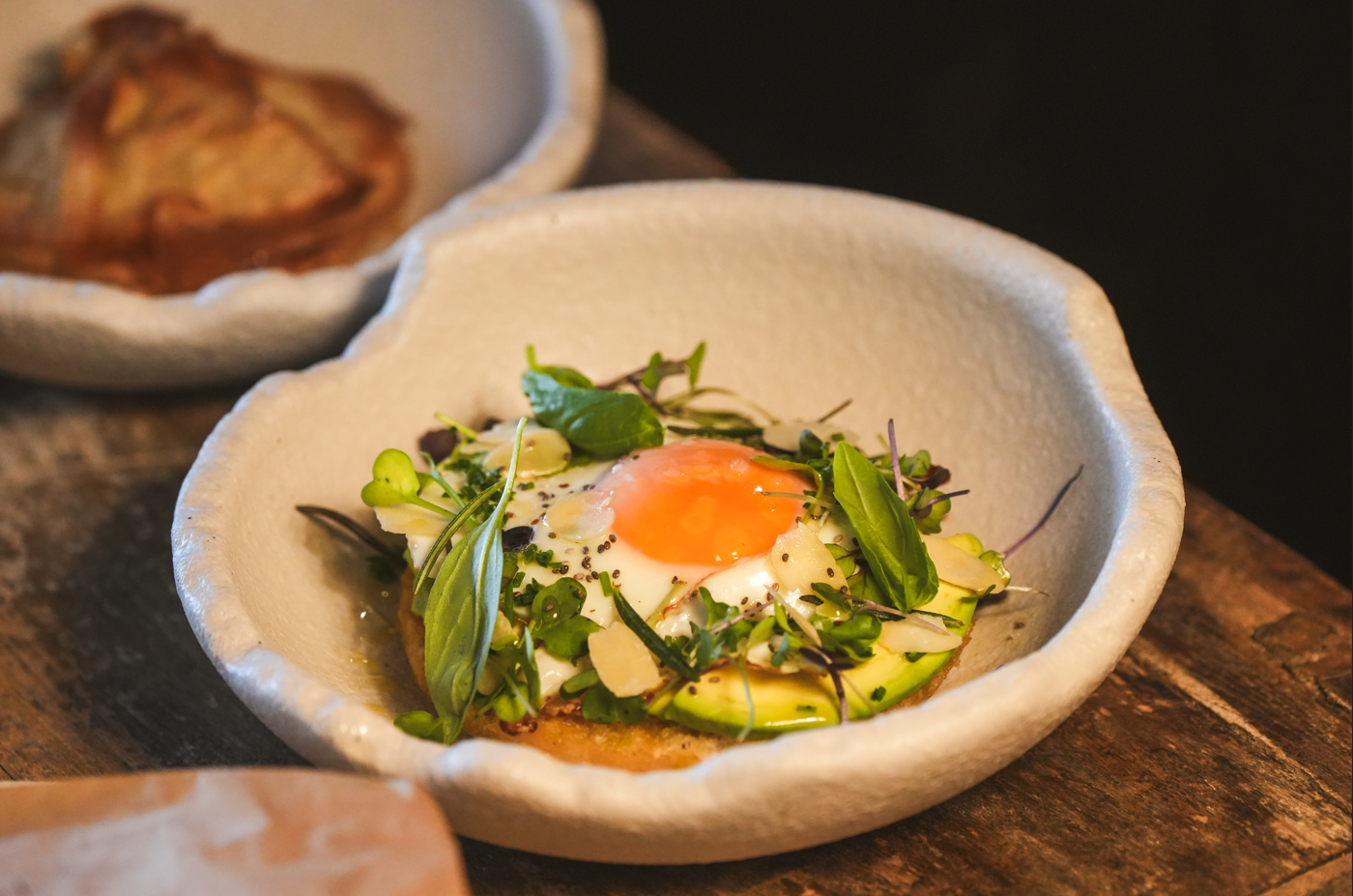 Un plato de brunch con un huevo frito, aguacate y verduras frescas se presenta en un cuenco blanco irregular sobre una mesa de madera.