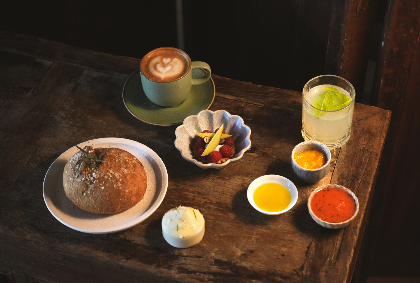 Una mesa de madera rústica exhibe una variedad de alimentos y bebidas, que incluyen café con leche, pan, un postre con frutos rojos, una bebida refrescante y varias salsas.