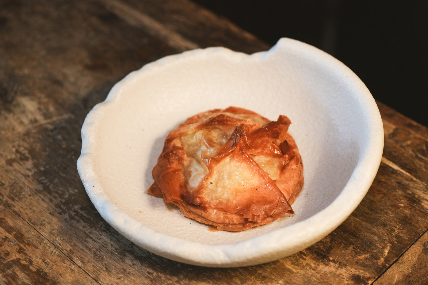 Un pastelito dorado de hojaldre se presenta en un cuenco blanco de borde irregular sobre una mesa de madera rústica.