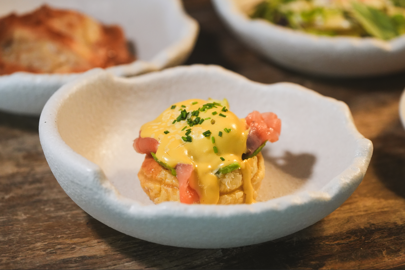 Un pequeño aperitivo de salmón, aguacate y salsa amarilla, adornado con cebollino, se sirve en un cuenco blanco de forma irregular sobre una mesa de madera.