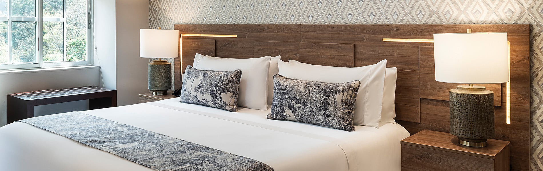 Dormitorio con papel tapiz de palmeras, cama king, mesita de noche y banco. Porton de la Cabrera