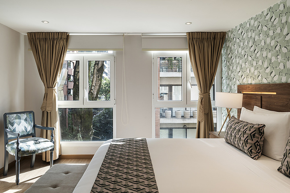 Habitación de hotel con cama grande, papel tapiz de grullas, ventana y silla decorativa. Porton de la Cabrera