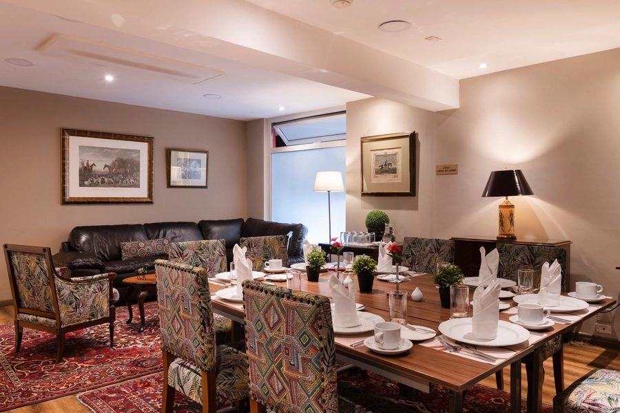 Salón Inglés del hotel Portón de la Cabrera con mesa de comedor montada, sillas tapizadas, sofá de cuero y alfombra persa.