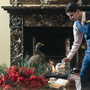 Un camarero sirve té junto a una chimenea, con un adorno de pavo real y flores rojas. ¡ElPorton de la Cabrera perfecto para tu estancia!