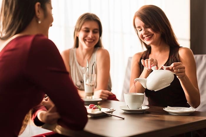 tres mujeres están sentadas en una mesa y una está vertiendo té en una taza .