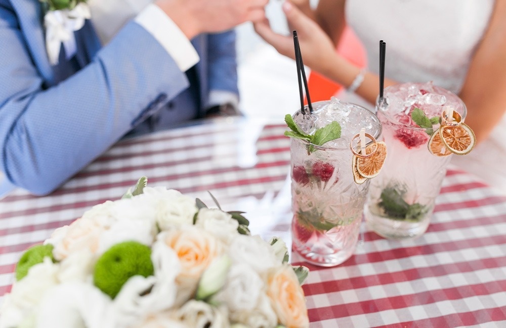 une mariée et un marié sont assis à une table avec deux cocktails à côté d' un bouquet de fleurs