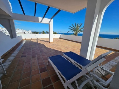 Amplia terraza con suelo de baldosas y dos tumbonas azules, ofreciendo vistas al mar y a la costa bajo un cielo despejado.