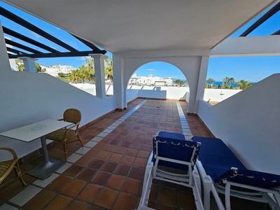 Una amplia terraza soleada con techo de pérgola, suelo de baldosas y muebles de exterior, ofrece vistas al mar, palmeras y edificios blancos bajo un cielo azul.