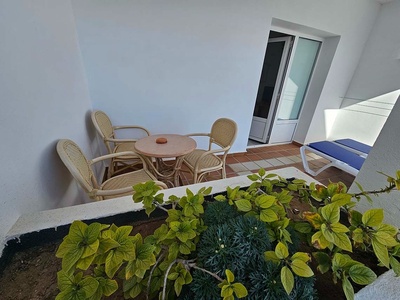 Un patio exterior con una mesa y sillas de mimbre, una tumbona y plantas en primer plano, con una puerta corredera al fondo.