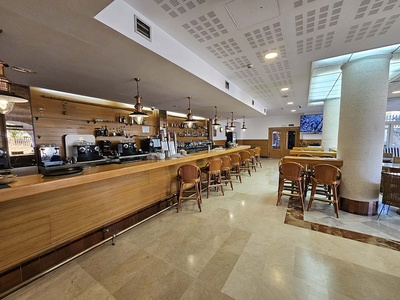 Un elegante bar interior se muestra con una larga barra de madera, taburetes de mimbre, máquinas de café, un suelo de mármol claro y una gran columna decorativa a la derecha. - 