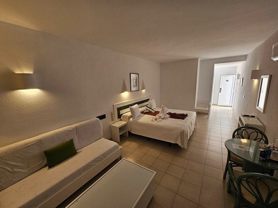 Se observa una espaciosa habitación de hotel o apartamento equipada con una cama, un sofá, una mesa con sillas y un estilo de decoración funcional de tonos claros.