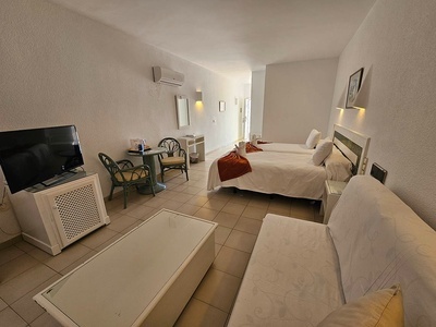 Una habitación de hotel sencilla y luminosa con dos camas individuales, un sofá, un televisor y una pequeña mesa con sillas.