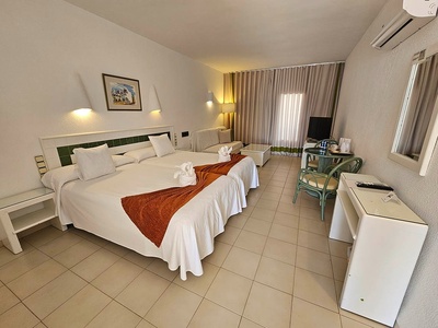 Una habitación de hotel sencilla y luminosa con dos camas individuales, un área de estar con sofá, televisión y una mesa con sillas.