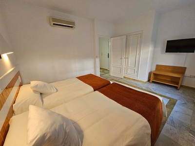 Una habitación de hotel sencilla con dos camas individuales, aire acondicionado, televisión y un banco de madera sobre un suelo de mármol.