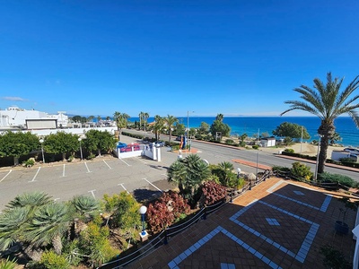 Una vista escénica de una zona costera con un aparcamiento, una carretera, vegetación exuberante y el mar azul brillante bajo un cielo despejado.