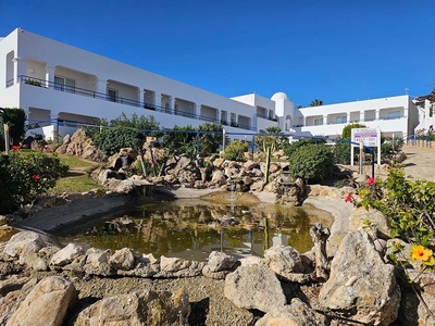 La imagen muestra un hotel blanco de estilo mediterráneo con balcones, rodeado de un jardín rocoso con un estanque, pequeñas cascadas y vegetación, bajo un cielo azul claro.