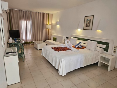 Una luminosa habitación de hotel con una cama doble adornada con toallas de cisne, una zona de estar con sofá y mesa, y un televisor.