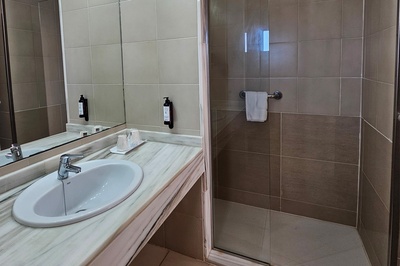 Un baño de tonos neutros con lavabo, encimera de mármol, un gran espejo, dispensadores de pared y una ducha con mampara de cristal.