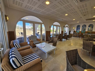 Un luminoso salón interior, posiblemente de un hotel, con muebles de mimbre, cojines a rayas azules y blancas, y grandes puertas acristaladas arqueadas que dan a un espacio exterior. - 