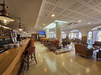 El interior de un bar o zona de descanso presenta una barra de madera con taburetes, mesas y sillones de mimbre, y arcos que conectan con el exterior. - 