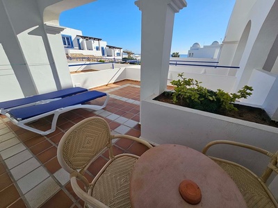 Una terraza soleada con mobiliario de exterior y plantas, enmarcada por arquitectura blanca y azul de estilo mediterráneo, con vista al mar bajo un cielo despejado.