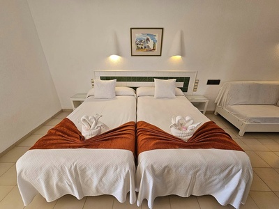 La imagen muestra una habitación de hotel con dos camas individuales decoradas con toallas en forma de cisne y mantas naranjas, un cabecero verde y un sofá-cama a la derecha.