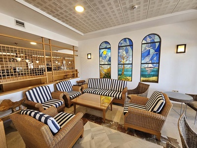 Una luminosa sala de estar presenta asientos de mimbre con cojines a rayas, tres vidrieras arqueadas de temática marina y una pared divisoria de celosía de madera. - 