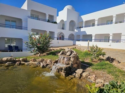 La imagen muestra un complejo de edificios blancos con barandales azules, un estanque con una cascada y jardines, bajo un cielo azul despejado.