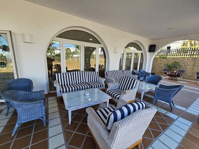 Un patio cubierto con mobiliario de mimbre, cojines a rayas azules y blancas, suelo de baldosas y arcos arquitectónicos. - 