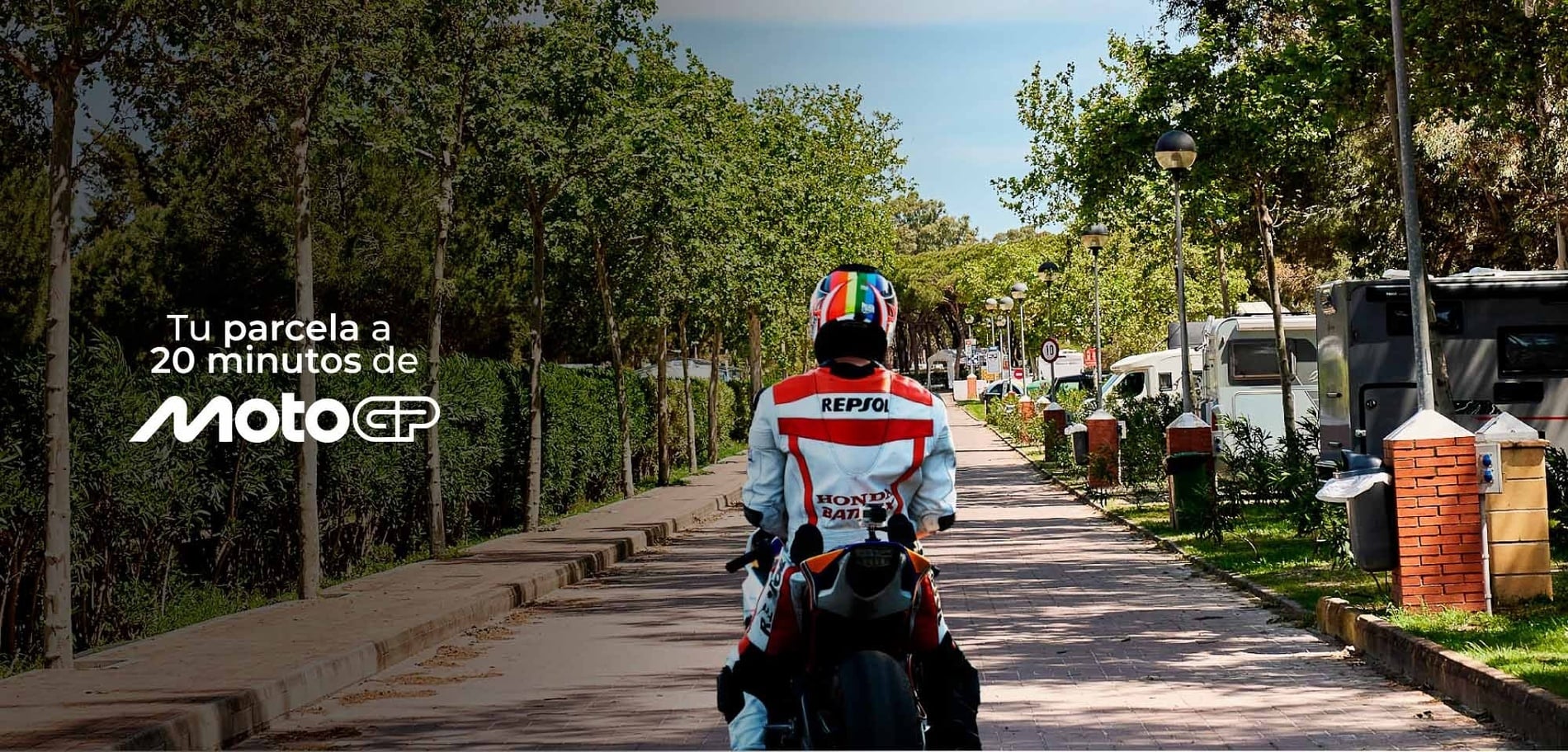Un motociclista con traje de competición transita por un camino arbolado en un entorno de camping con caravanas, junto al texto "Tu parcela a 20 minutos de MotoGP".
