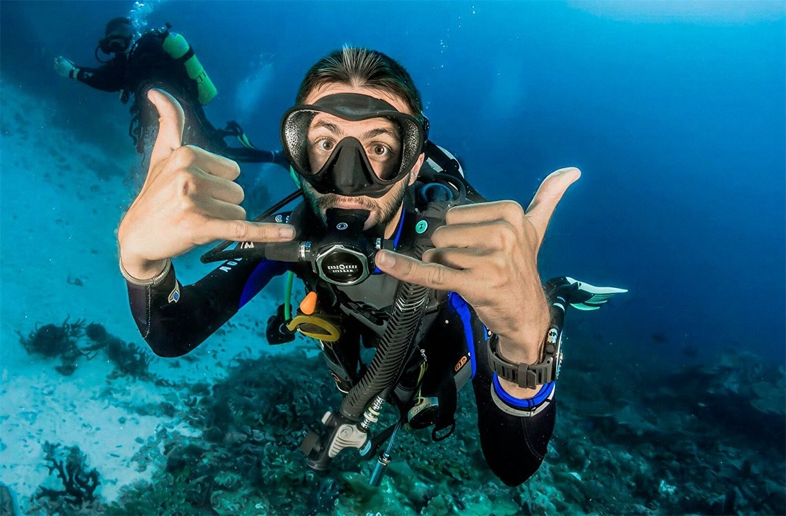 Un buzo masculino con equipo de buceo mira a la cámara con los ojos muy abiertos y hace el signo shaka con ambas manos, mientras otro buzo se ve en el fondo.