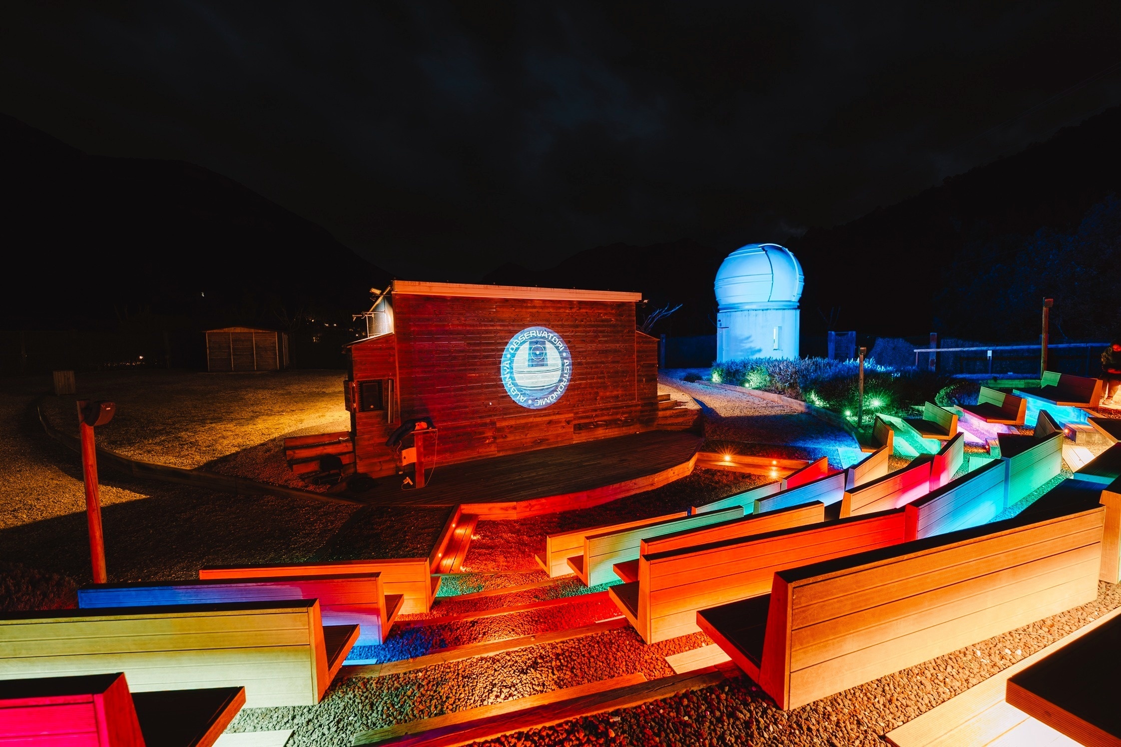 Observatorio astronómico nocturno con anfiteatro exterior y luces de colores.